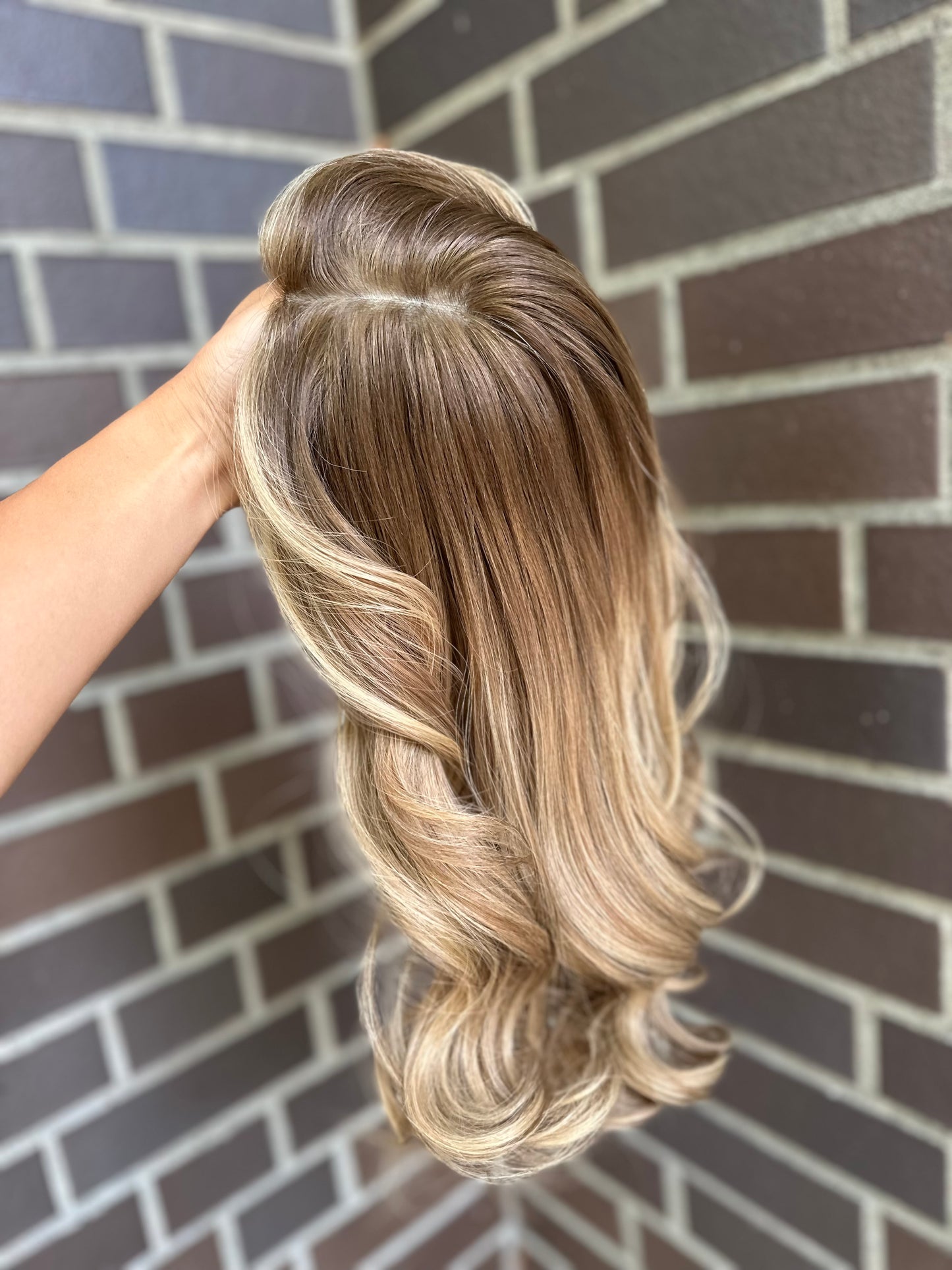Bild zeigt blondes Echthaarteil, das seitlich ins Bild gehalten wird. Bild dient der Produktpräsentation eines Artikels in der Kategorie Topper/Perücken.