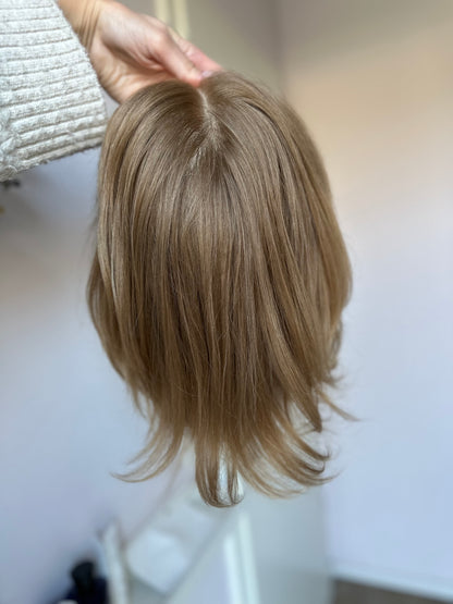 Bild zeigt blondes Echthaarteil, das frontal ins Bild gehalten wird. Bild dient der Produktpräsentation eines Artikels in der Kategorie Topper/Perücken.