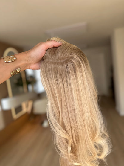 Bild zeigt blondes Echthaarteil, das seitlich ins Bild gehalten wird. Bild dient der Produktpräsentation eines Artikels in der Kategorie Topper/Perücken.