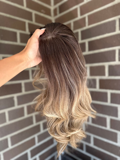 Bild zeigt blondes Echthaarteil, das seitlich ins Bild gehalten wird. Bild dient der Produktpräsentation eines Artikels in der Kategorie Topper/Perücken.