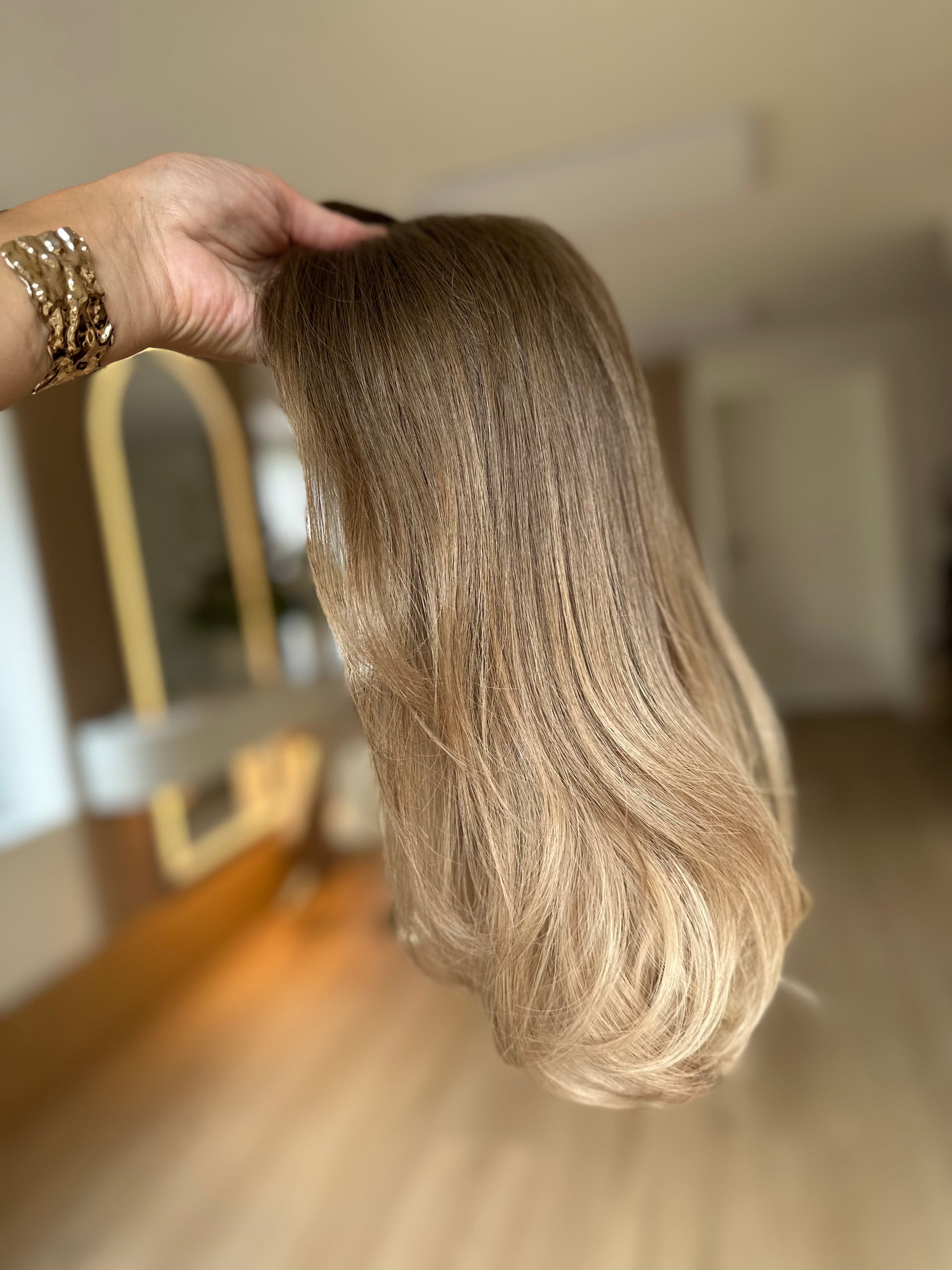 Bild zeigt blondes Echthaarteil, das seitlich ins Bild gehalten wird. Bild dient der Produktpräsentation eines Artikels in der Kategorie Topper/Perücken.
