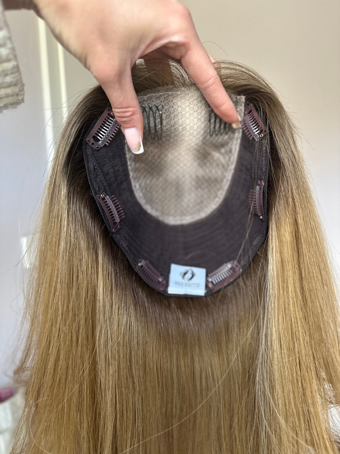 Bild zeigt blondes Echthaarteil, das frontal ins Bild gehalten wird. Bild dient der Produktpräsentation eines Artikels in der Kategorie Topper/Perücken.