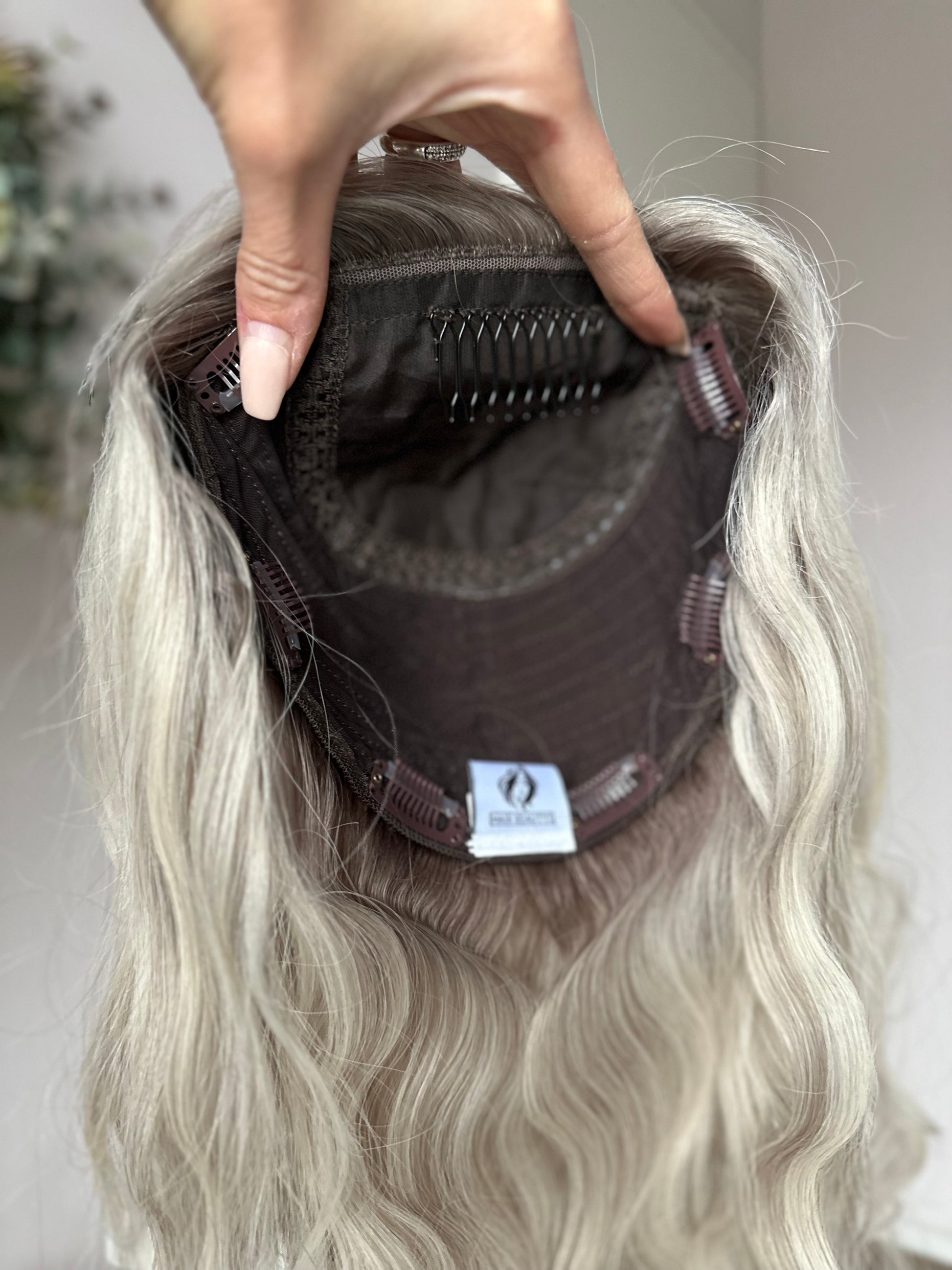 Bild zeigt blondes Echthaarteil, das frontal ins Bild gehalten wird. Bild dient der Produktpräsentation eines Artikels in der Kategorie Topper/Perücken.