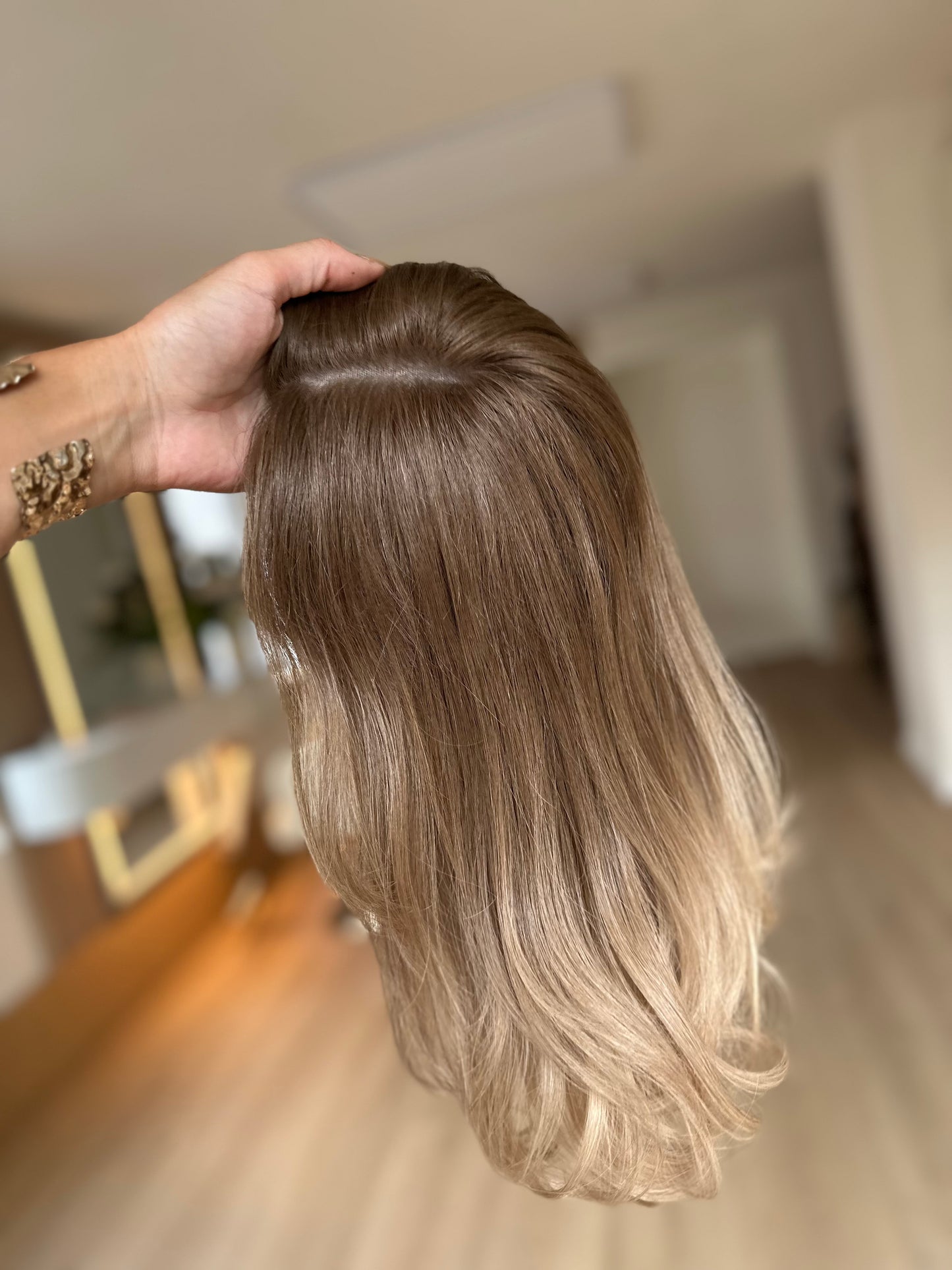 Bild zeigt blondes Echthaarteil, das seitlich ins Bild gehalten wird. Bild dient der Produktpräsentation eines Artikels in der Kategorie Topper/Perücken.
