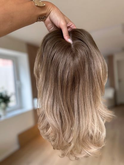Bild zeigt blondes Echthaarteil, das frontal ins Bild gehalten wird. Bild dient der Produktpräsentation eines Artikels in der Kategorie Topper/Perücken.